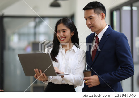 Two Asian businesspeople are standing in office, collaborating on a project using a laptop, teamwork and corporate. Two Asian businesspeople are standing in office, collaborating on a project using a laptop, teamwork and corporate. 133947340