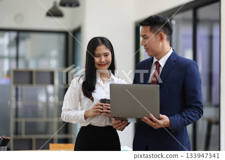 Two Asian businesspeople are standing in office, collaborating on a project using a laptop, teamwork and corporate. Two Asian businesspeople are standing in office, collaborating on a project using a laptop, teamwork and corporate. 133947341