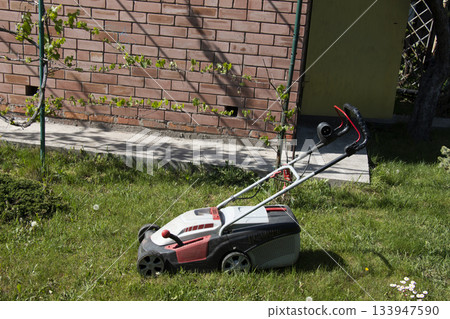 Side view of a modern electric lawn mower cutting bright, lush green grass. Garden tools. Rotary lawn mower on the lawn. Professional lawn care. Side view of a modern electric lawn mower cutting bright, lush green grass. Garden tools. Rotary lawn mower on the lawn. Professional lawn care. 133947590
