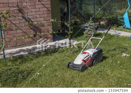 A side view of a modern electric lawn mower cutting bright, lush green grass. Garden tools. Rotary lawn mower on the lawn. Professional lawn care. 133947594