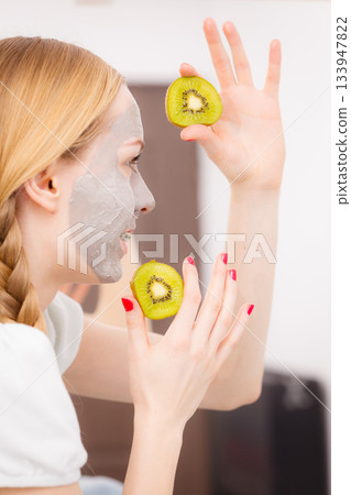 Happy young woman having face mask holding kiwi Happy young woman having face mask holding kiwi 133947822