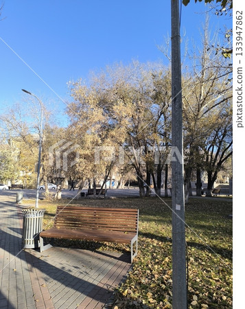 A peaceful park bench surrounded by trees with golden autumn leaves. The clear blue sky complements the scene, while fallen leaves on the grass enhance the seasonal charm. 133947862