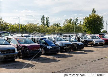 Rows of dismantled cars stand on open yard, showing missing parts, broken fronts exposed interiors. Scene reflects scrapyard atmosphere scattered vehicles, warm sunlight and industrial surroundings Rows of dismantled cars stand on open yard, showing missing parts, broken fronts exposed interiors. Scene reflects scrapyard atmosphere scattered vehicles, warm sunlight and industrial surroundings 133948003