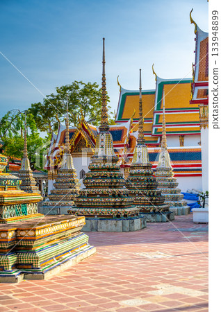 Exterior view of Wat Pho with richly decorated stupas covered in colorful floral ceramics and golden temple roofs shining under a clear blue sky on a bright sunny day in Bangkok. 133948899