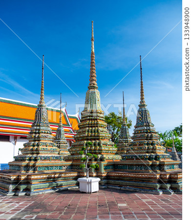 Exterior view of Wat Pho with richly decorated stupas covered in colorful floral ceramics and golden temple roofs shining under a clear blue sky on a bright sunny day in Bangkok. 133948900