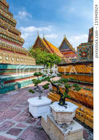 Exterior view of Wat Pho with richly decorated stupas covered in colorful floral ceramics and golden temple roofs shining under a clear blue sky on a bright sunny day in Bangkok. 133948924