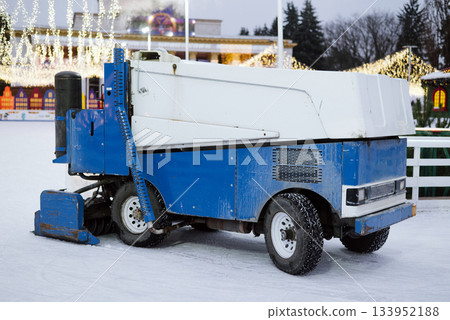 Ice resurfacer machine in blue and white smoothing the surface of an outdoor skating rink decorated with bright holiday lights. 133952188