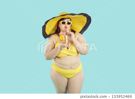 Carefree young plus size woman wearing beachwear drinking cocktail on blue studio background. 133952469