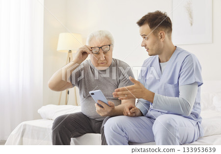 Young cheerful male nurse showing elderly man how to use smartphone in clinic Young cheerful male nurse showing elderly man how to use smartphone in clinic 133953639
