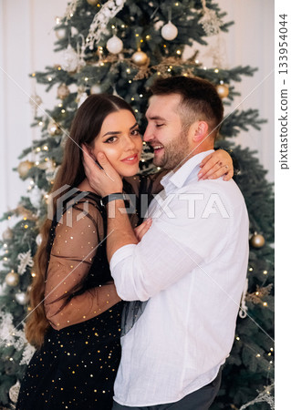 Couple embraces in festive attire by decorated Christmas tree during holiday celebration in cozy indoor setting 133954044