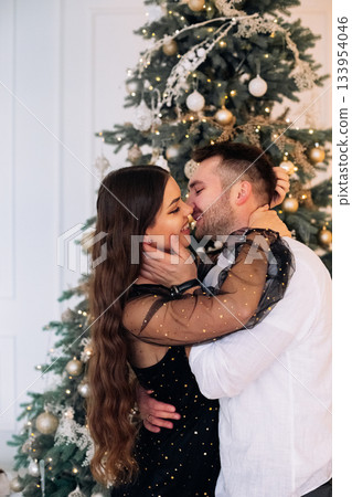 Couple embraces joyfully by a beautifully decorated Christmas tree indoors, celebrating love and togetherness during the holiday season Couple embraces joyfully by a beautifully decorated Christmas tree indoors, celebrating love and togetherness during the holiday season 133954046