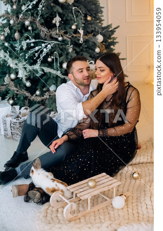Couple enjoying a cozy moment by a beautifully decorated Christmas tree with their rabbit in a festive home setting during the holiday season Couple enjoying a cozy moment by a beautifully decorated Christmas tree with their rabbit in a festive home setting during the holiday season 133954059