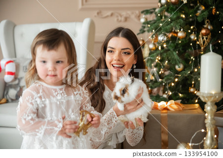 Happy child and adult celebrating holiday season with bunny near Christmas tree in cozy living room filled with decorations and gifts 133954100