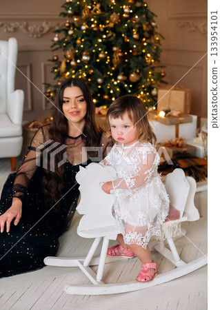 Child enjoys rocking horse while adult sits nearby in a cozy holiday setting decorated with a glowing Christmas tree and gifts 133954101