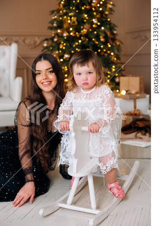 Young girl in a beautiful dress plays on a rocking horse with her mother nearby, festive decorations adding warmth to the room during the holiday season Young girl in a beautiful dress plays on a rocking horse with her mother nearby, festive decorations adding warmth to the room during the holiday season 133954112