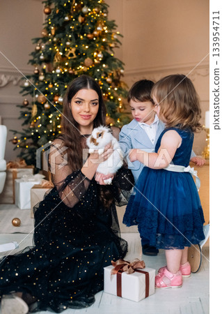 Family celebration with children and a pet rabbit during holiday season, surrounded by gifts and Christmas tree in a cozy indoor setting 133954711