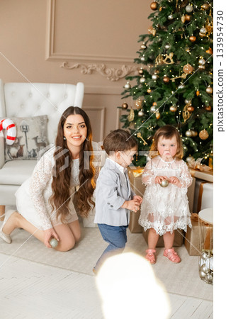 Children playing with ornaments while a woman supervises and smiles, embracing the festive spirit during a holiday celebration in a beautifully decorated living room 133954730