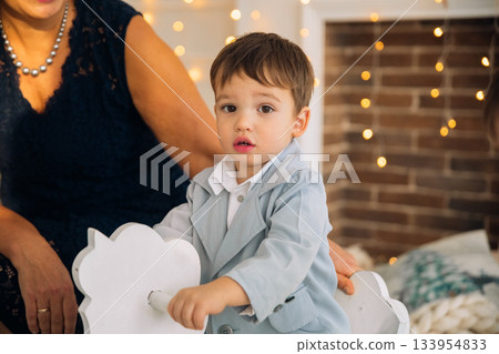 Child riding on rocking horse while being watched by adult in a cozy, decorated indoor space with fairy lights, creating a warm atmosphere in the evening Child riding on rocking horse while being watched by adult in a cozy, decorated indoor space with fairy lights, creating a warm atmosphere in the evening 133954833