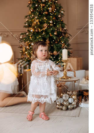 Toddler in a white dress stands near a beautifully decorated Christmas tree surrounded by gifts, capturing a festive and joyful moment 133954838