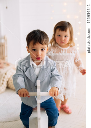 Children playing together in a cozy indoor space, one riding a toy while the other watches with curiosity 133954841