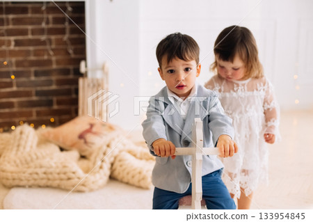 Children playing indoors during a cozy moment, a boy riding a toy while a girl observes, creating a warm atmosphere in a sunny room Children playing indoors during a cozy moment, a boy riding a toy while a girl observes, creating a warm atmosphere in a sunny room 133954845