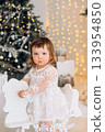 Charming toddler in a white dress plays on a wooden rocking horse near a beautifully decorated Christmas tree with fairy lights 133954850
