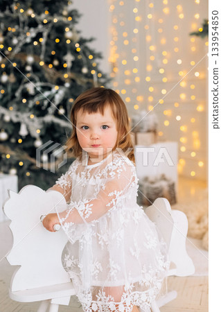 Charming toddler in a white dress plays on a wooden rocking horse near a beautifully decorated Christmas tree with fairy lights Charming toddler in a white dress plays on a wooden rocking horse near a beautifully decorated Christmas tree with fairy lights 133954850