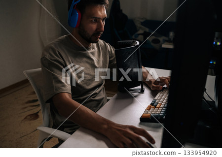 Gamer wearing colorful headphones playing computer video games at desktop setup, streaming online content and participating in esports competition from home or internet cafe. Gamer wearing colorful headphones playing computer video games at desktop setup, streaming online content and participating in esports competition from home or internet cafe. 133954920