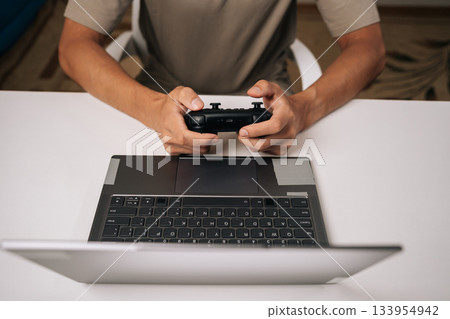 Top view of gamer male gaming on laptop computer using black wireless gamepad controller, enjoying modern online entertainment during leisure time, hands holding joystick device over keyboard. Top view of gamer male gaming on laptop computer using black wireless gamepad controller, enjoying modern online entertainment during leisure time, hands holding joystick device over keyboard. 133954942