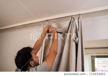 Man reaching up to attach light-colored curtain to white track rod on ceiling, focusing on setting up home decor in apartment, representing domestic life. Concept of household cleanliness. Man reaching up to attach light-colored curtain to white track rod on ceiling, focusing on setting up home decor in apartment, representing domestic life. Concept of household cleanliness. 133954943