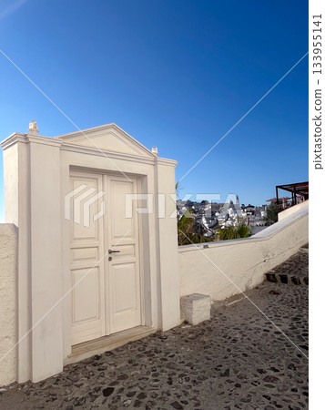 Greek island scene with a classic white door opening onto a cobblestone street, offering a picturesque village view and the Aegean sea beyond under a clear blue sky. Greek island scene with a classic white door opening onto a cobblestone street, offering a picturesque village view and the Aegean sea beyond under a clear blue sky. 133955141