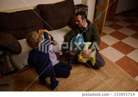A father sits on the floor with gift bags while his two children play nearby in a cozy home Christmas scene. 133955714