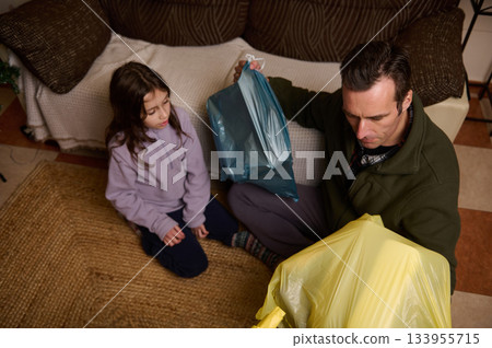 A man and a young girl sit on a rug in a cozy living room, handling colorful plastic bags, signaling a moment of family shopping or cleanup at home. 133955715