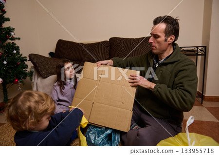 A father sits on the floor with a large cardboard box as two children nearby prepare gifts. A Christmas tree adds festive warmth to this cozy family moment. A father sits on the floor with a large cardboard box as two children nearby prepare gifts. A Christmas tree adds festive warmth to this cozy family moment. 133955718