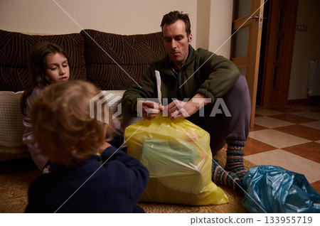 A father sits on the floor with two children, packing household trash into colorful bags. They focus on tidying up a cozy living room, turning a routine chore into a family moment. A father sits on the floor with two children, packing household trash into colorful bags. They focus on tidying up a cozy living room, turning a routine chore into a family moment. 133955719