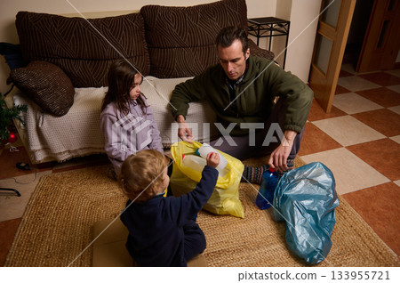 A father sits with two kids on the living room rug, packing items into yellow and blue bags during a holiday cleanup in a cozy home. 133955721