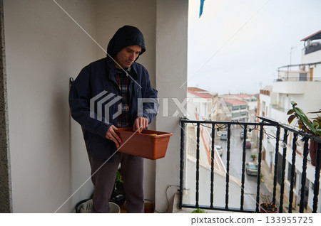 A man in a hooded jacket tends a terracotta planter on a balcony overlooking a city street. A calm, chilly urban moment focused on home gardening. A man in a hooded jacket tends a terracotta planter on a balcony overlooking a city street. A calm, chilly urban moment focused on home gardening. 133955725