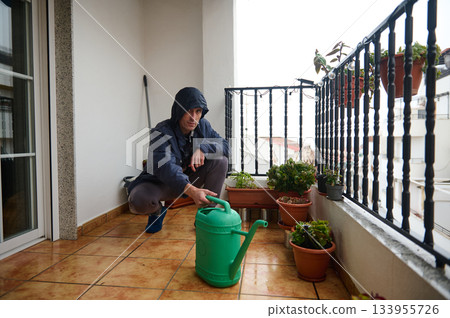 A man in a dark hoodie waters plants on a sunlit balcony, surrounded by potted greenery and a protective railing. 133955726