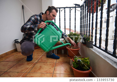 A man on a balcony waters potted plants with a bright green watering can. A casual, home garden moment showcasing plant care and outdoor living. 133955730