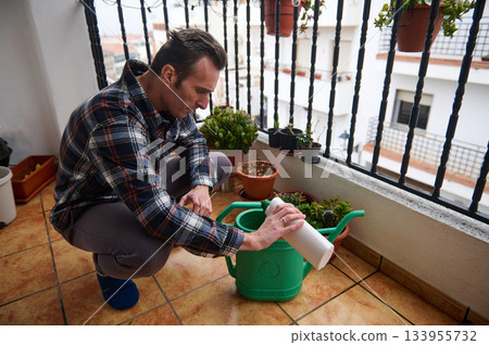 A man kneels on a balcony, tending potted plants. He uses a spray bottle to water and nurture his small garden, surrounded by pots and railing in an urban setting. A man kneels on a balcony, tending potted plants. He uses a spray bottle to water and nurture his small garden, surrounded by pots and railing in an urban setting. 133955732