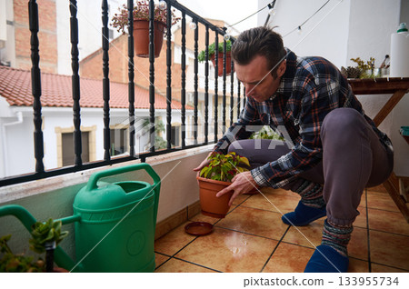 A man kneels on a tiled balcony, tending a potted plant. A green watering can sits nearby, with string lights creating a warm urban garden scene. 133955734