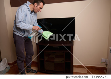 A man in casual wear cleans a flat-screen TV with a spray bottle and microfiber cloth in a cozy living room, highlighting home maintenance and everyday household chores. A man in casual wear cleans a flat-screen TV with a spray bottle and microfiber cloth in a cozy living room, highlighting home maintenance and everyday household chores. 133955740