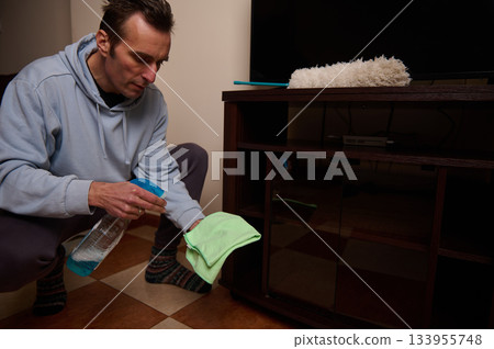 A man in a light blue hoodie cleans a television stand with a spray bottle and soft cloth, maintaining a neat living room and home care routine. A man in a light blue hoodie cleans a television stand with a spray bottle and soft cloth, maintaining a neat living room and home care routine. 133955748