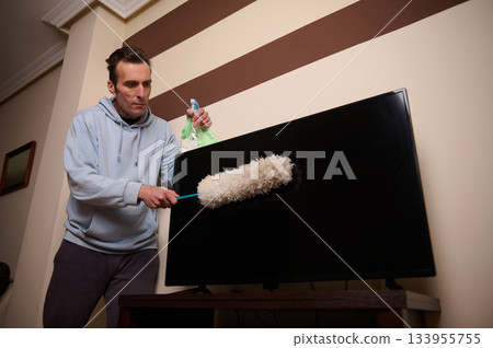 A man in a light blue hoodie uses a paint roller to clean a large flat-screen television, holding a spray bottle, in a cozy living room with striped wall decor. 133955755