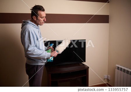 A man in a light hoodie cleans a flat screen TV using a spray bottle and fluffy duster in a cozy living room. Clean, home maintenance, and everyday chores captured candidly. A man in a light hoodie cleans a flat screen TV using a spray bottle and fluffy duster in a cozy living room. Clean, home maintenance, and everyday chores captured candidly. 133955757