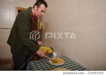 A man in a green jacket serves potato wedges and noodles on a checkered tablecloth in a simple tiled kitchen, creating a casual, homely meal scene. A man in a green jacket serves potato wedges and noodles on a checkered tablecloth in a simple tiled kitchen, creating a casual, homely meal scene. 133955763