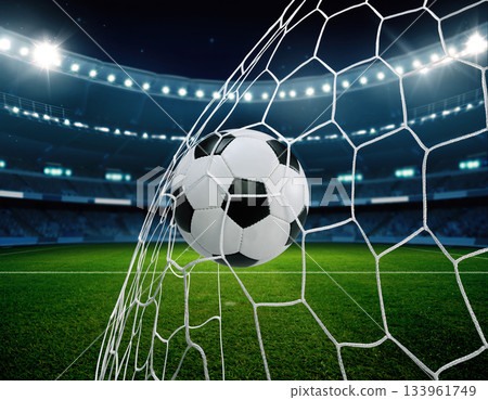 Soccer ball hitting the net in a stadium during a match at night with bright lights shining down 133961749