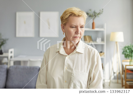 Pensive elderly woman standing alone in living room, looking thinking, thoughtful mature wife 133962317