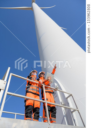 Two engineers in safety uniform inspecting wind turbine. Male worker holding laptop while female communicates via radio walkie talkie while standing on platform Two engineers in safety uniform inspecting wind turbine. Male worker holding laptop while female communicates via radio walkie talkie while standing on platform 133962808