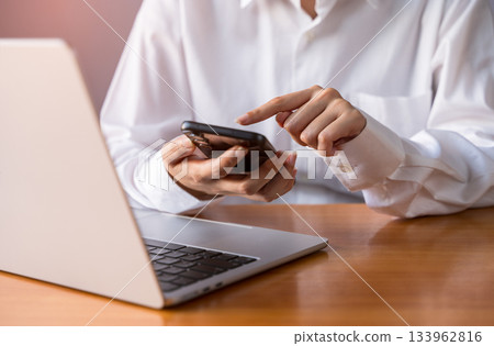 Woman in white shirt using smartphone while sitting at office wooden desk with laptop Woman in white shirt using smartphone while sitting at office wooden desk with laptop 133962816